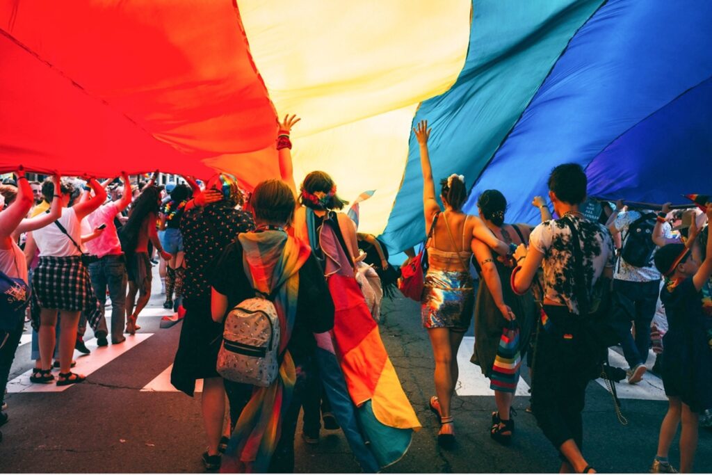 Un grupo de personas camina bajo una gran bandera arco iris durante una Marcha del Orgullo