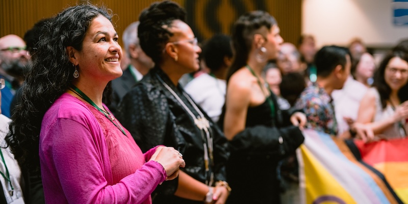 A group of diverse individuals gathered at an event, with a focus on one person in the foreground wearing a bright pink top and smiling warmly. Behind them, others are engaged in the event, some holding the intersex-inclusive Pride flag. The atmosphere appears lively and inclusive, with people of diverse styles and expressions present in the background.