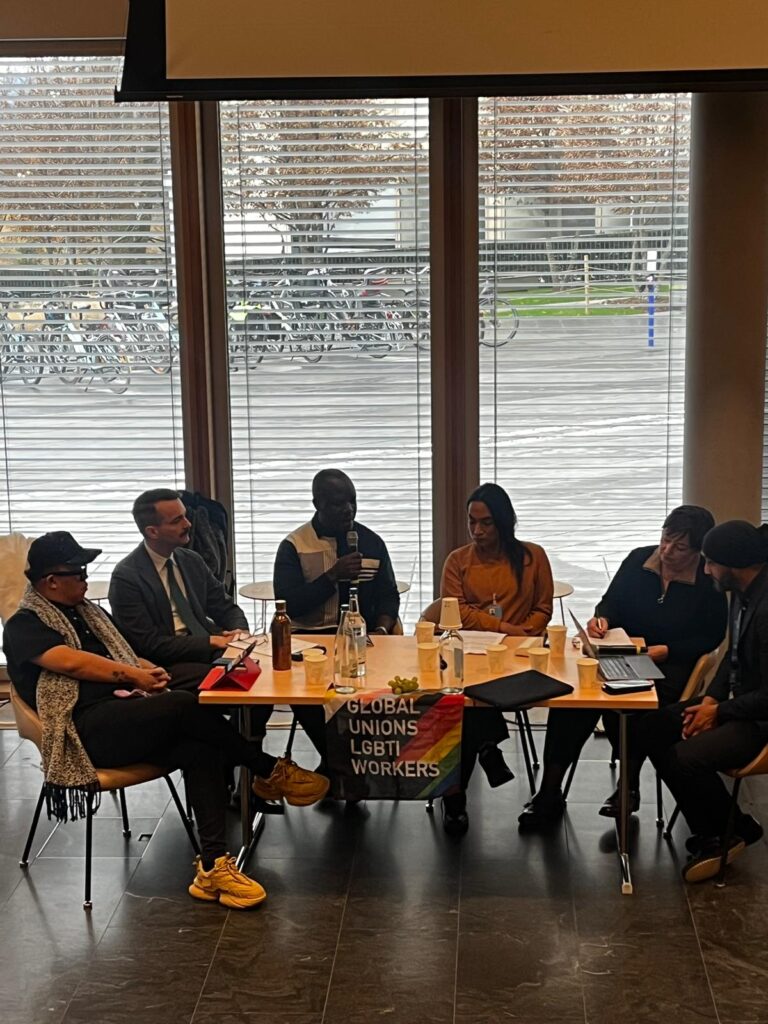 A group of diverse people sitting at a panel organized by ILGA World in front of the reception of building H l at the Palais de Nations in Geneva, Switzerland. 