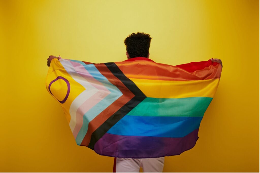 A person is turning their back to the camera and holds an intersex-inclusive Progress Pride flag