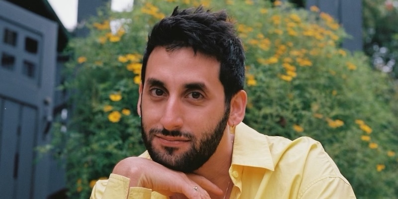 A close-up of a person with dark hair and a beard, wearing a yellow short, and posing outdoors in front of a hedge full of yellow flowers