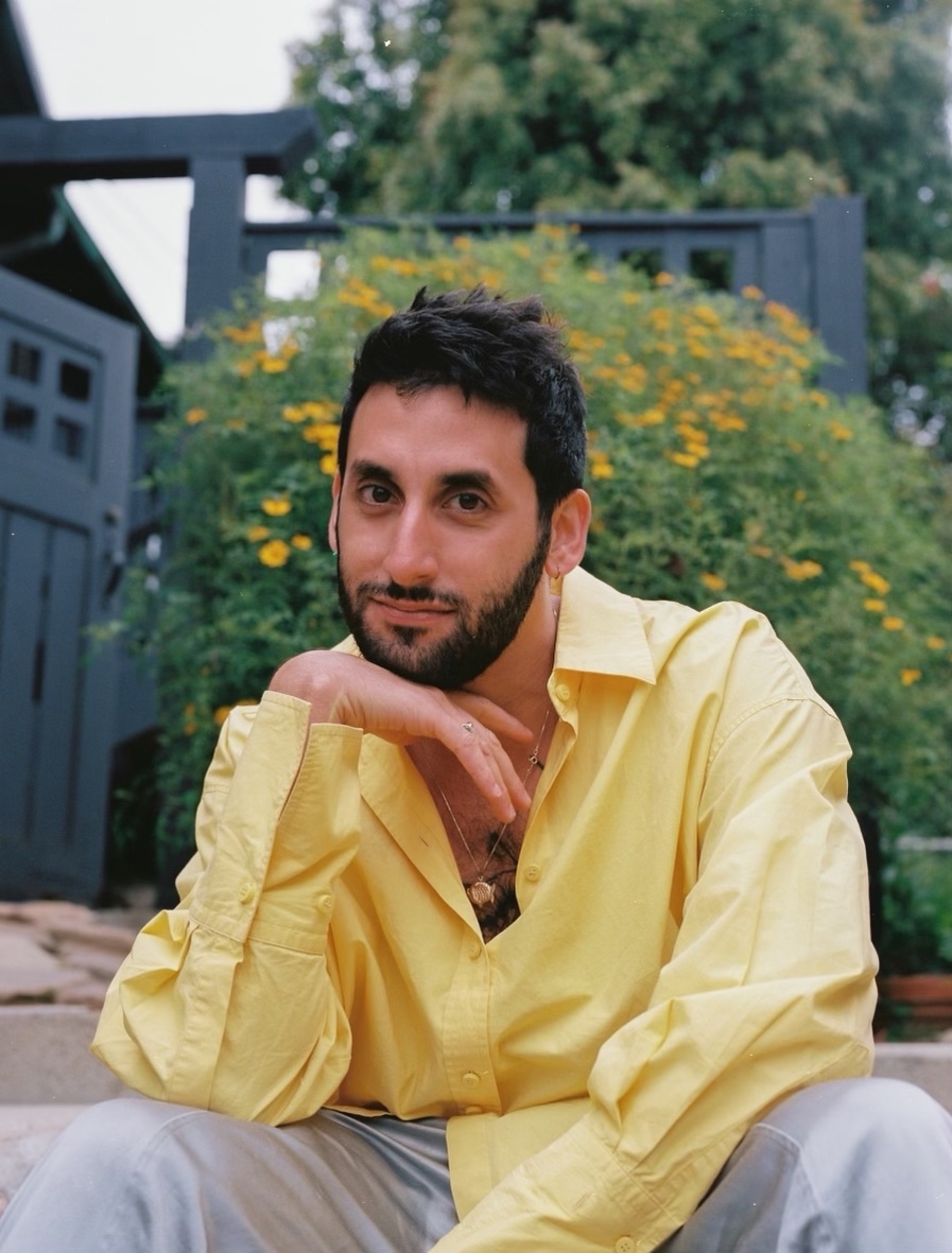 A portrait of a person with dark hair and a beard, wearing a yellow short, and posing outdoors in front of a hedge full of yellow flowers