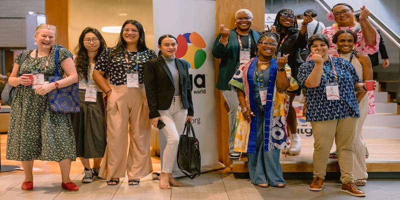 A group of diverse people smiles for the camera, posing in front of an ILGA World banner. Some of them make the "heart" gesture with their hands
