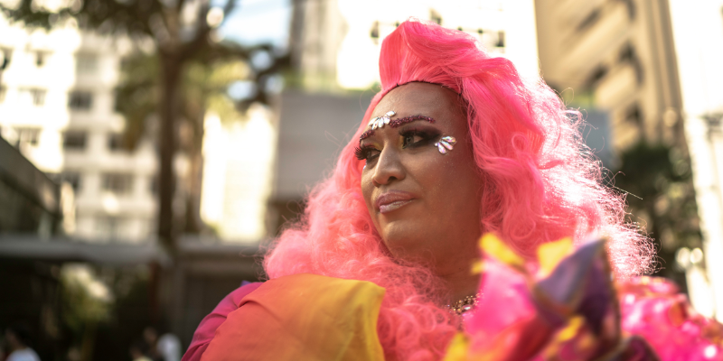 A drag artist with a pink wig looks away from the camera in a city setting